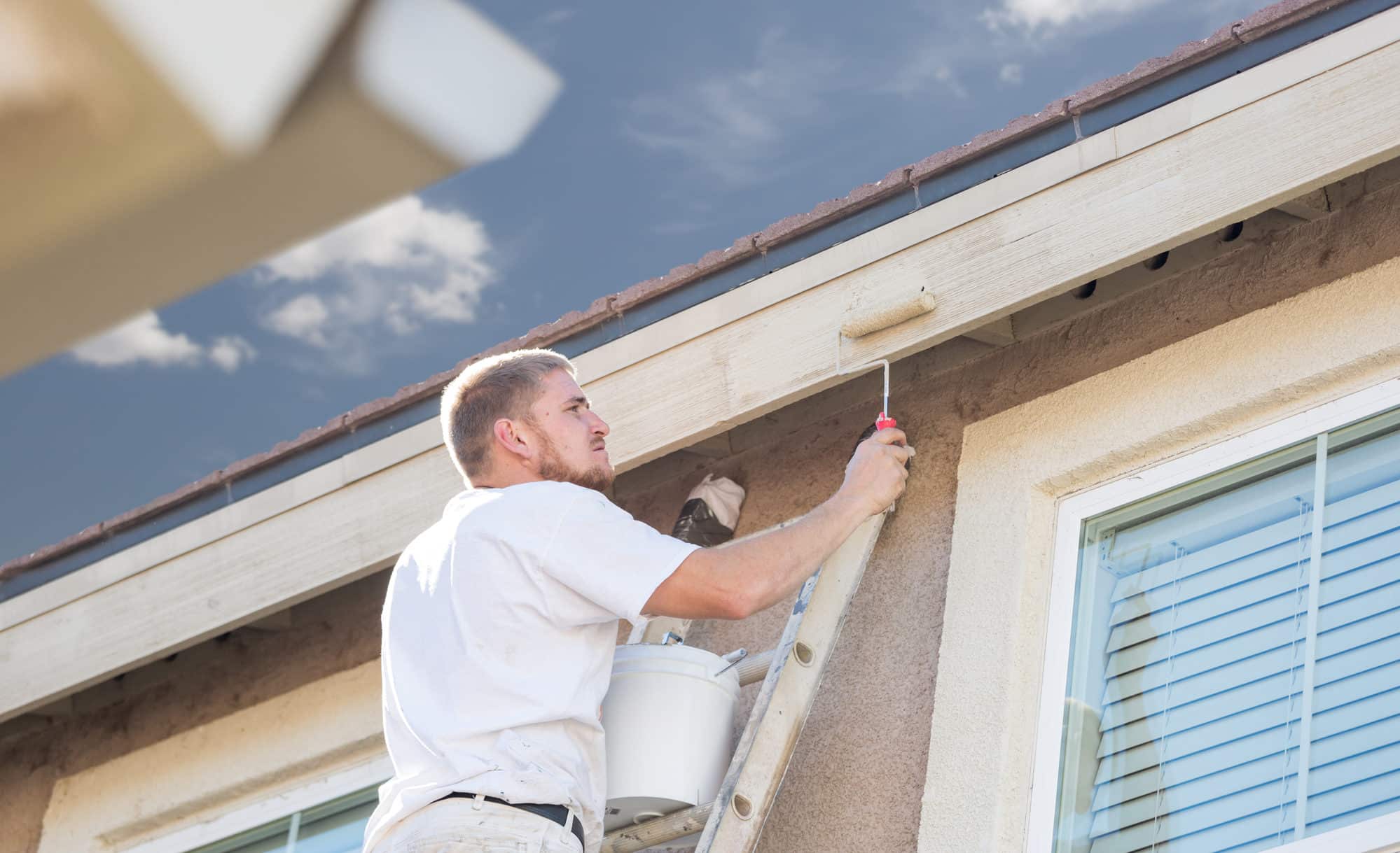 Painter on a ladder painting white fascia and eaves under a blue sky