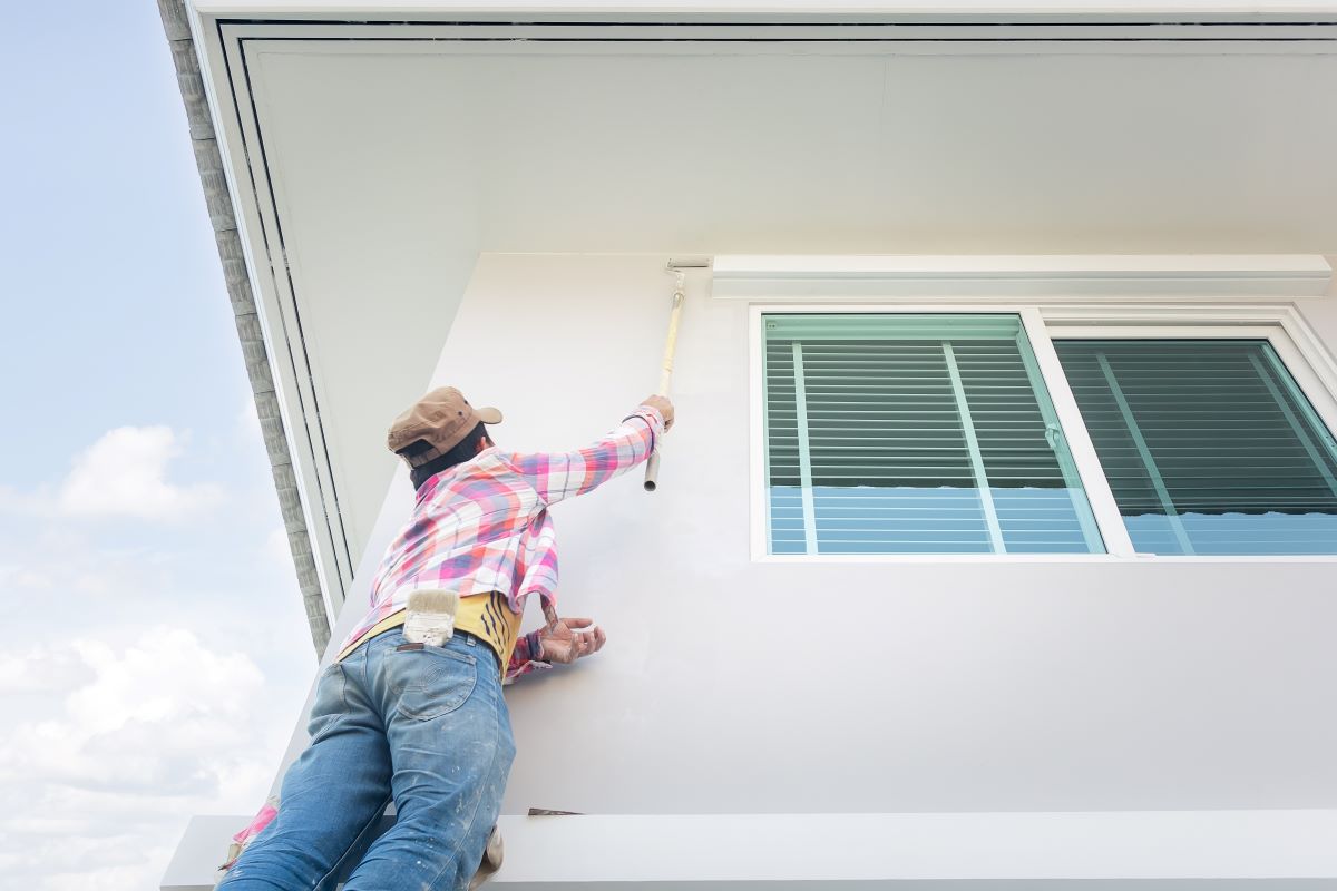 Painter on a ladder painting the exterior wall of a home beside a window